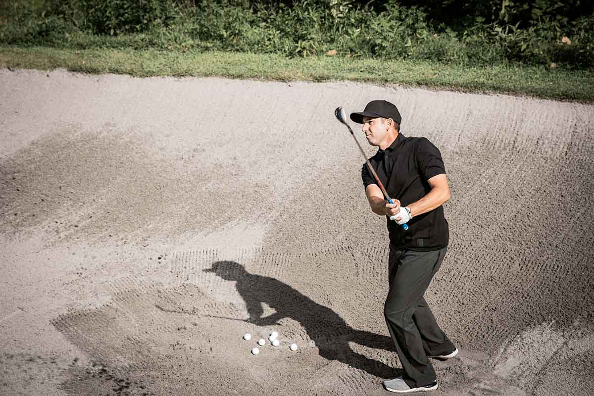 golfer swinging golf club while standing in a sand